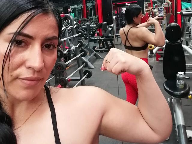 SofiaAnal hat dunkles Haar, eine kleine Nase und helle Haut. Sie zeigt einen angespannten Bizeps in einem schwarzen Sport-BH, roten Leggings und ein Bild von ihr im Fitnessstudio mit Hanteln..