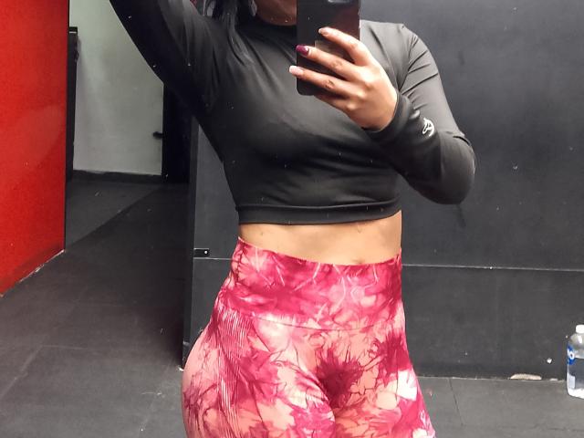 SofiaAnal, met een gemiddelde borstmaat, draagt een zwart shirt met lange mouwen en korte broekjes met een roze bloemenprint, en maakt een spiegelzelfportret in een sportschool..