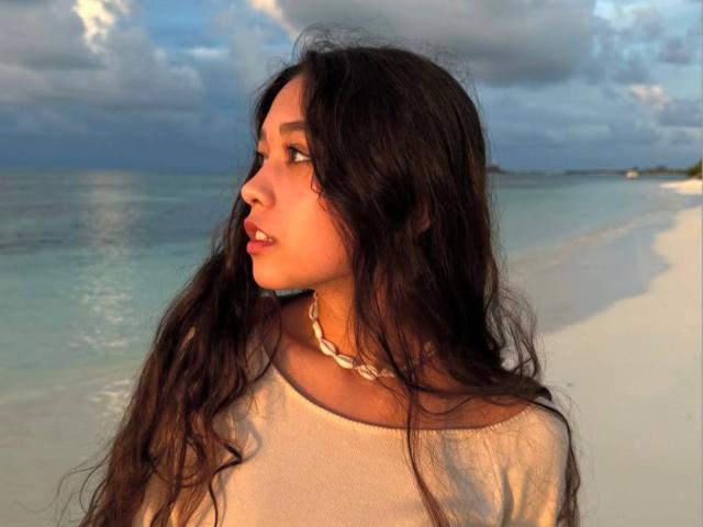 LourieSteins has long, wavy dark hair, a tan skin tone, and wears a white shirt with a shell necklace. She's looking to the side with a pouty expression.
