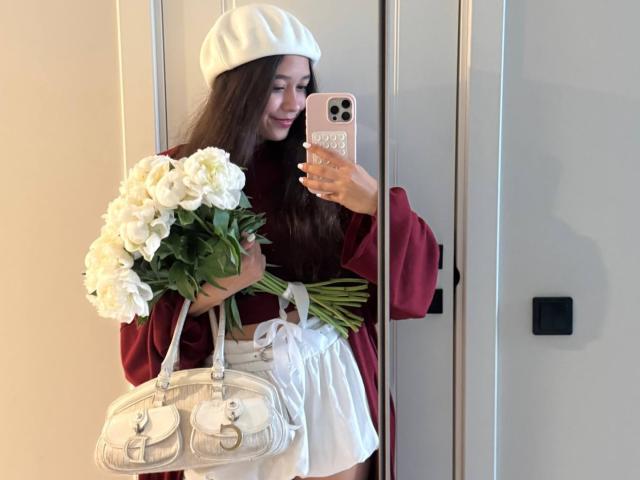 LourieSteins met lang bruin haar, een witte baret, een rode trui, een witte rok, die witte rozen en een zilveren handtas vasthoudt, en een spiegel-selfie maakt met een roze telefoon..