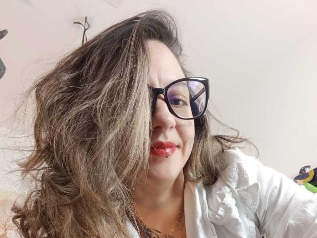 ArianaBlonde has wavy brown hair, black glasses, and red lipstick. She's wearing a white shirt, showing a bit of her chest, with a slightly smiling face.