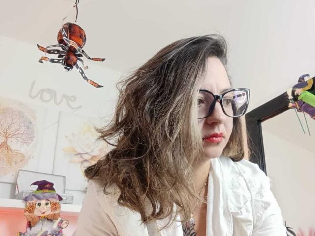 ArianaBlonde with wavy brown hair, glasses, and red lipstick, wearing a white top, looking at the camera, with a spider ornament and a small doll in the background.
