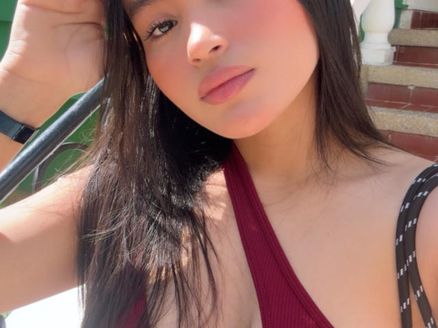 MiiaPalmer with long black hair, pink lips, wearing a deep red halter top, black strap dress, and black wristwatch, holding a black strap, sunny, soft light.