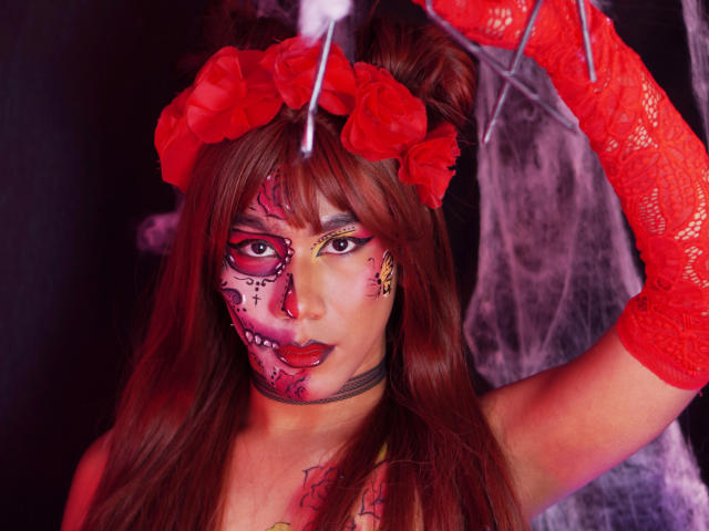 ScarlettHotSweet, avec ses cheveux bruns, des roses rouges dans un serre-tête, des gants en dentelle rouge, le visage maquillé comme un crâne de sucre, et des tatouages, lève le bras, exhibant sa poitrine et son anneau de nez..