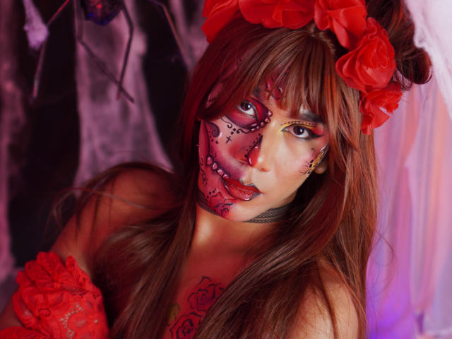 ScarlettHotSweet with red rose headpiece, face painted in sugar skull style, wearing red lace gloves, black choker, and tattoos, gazing at camera with purple eye makeup.