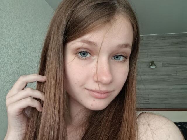 JessicaChrystal has light brown hair, blue eyes, and a small nose. She's touching her hair with a slight smile, showing a few small moles on her face.