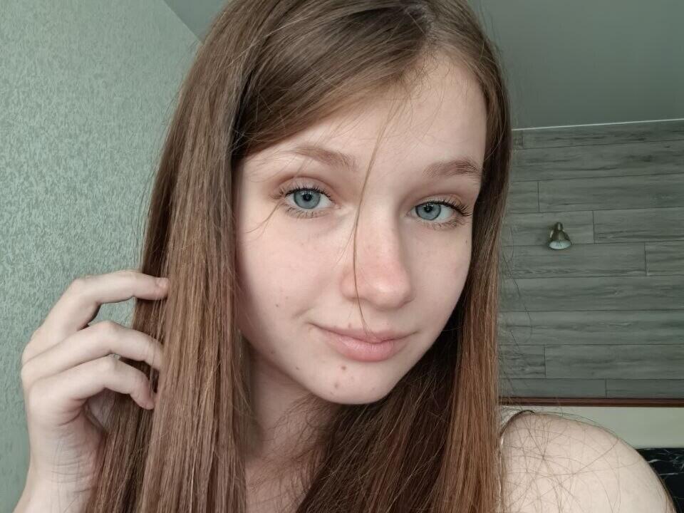 JessicaChrystal has light brown hair, blue eyes, and a small nose. She's touching her hair with a slight smile, showing a few small moles on her face.
