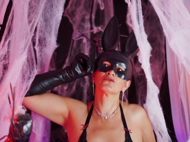 MilenneSexy, vêtue de gants en cuir noir, d'un masque avec des oreilles de lapin, d'un haut à encolure plongeante, d'un collier et de boucles d'oreilles cercles, a l'air sérieuse, avec un fond rose et noir. Seins visibles, bras levés, le masque couvre partiellement le visage..