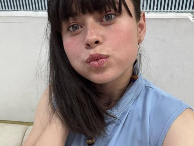 Iniridax with puffy pink lips, fair skin, and dark brown hair, wearing a sleeveless blue top, showing off small tits, and a beaded necklace, looking at the camera.