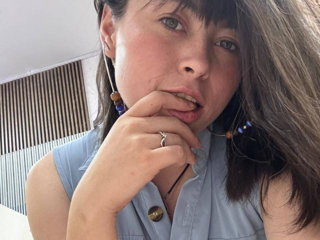 Iniridax with brown hair, blue eyes, and light skin, biting her finger, wearing a sleeveless light blue shirt, and a ring on her finger, looking slightly seductive.