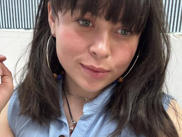 Iniridax has pale skin, green eyes, and dark brown hair with bangs. She's wearing a blue shirt, hoop earrings, and a necklace, with her tongue slightly out.