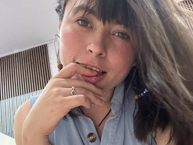 Iniridax with brown hair, green eyes, and a light blue shirt, biting her finger with a ring and necklace, looking slightly bored. Her tits are small and she's cute.