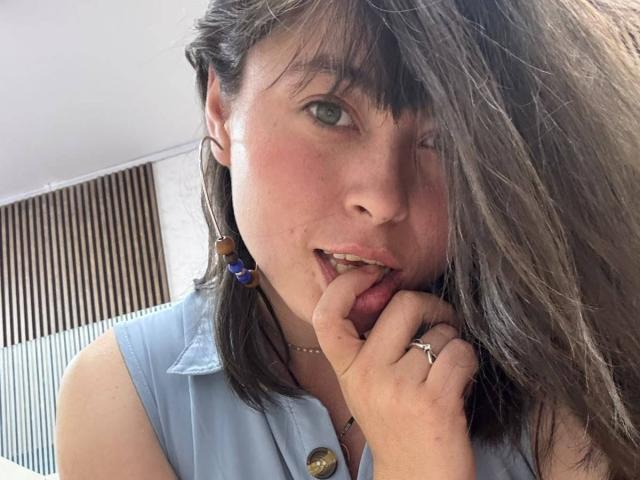 Iniridax with dirty brown hair, blue shirt, biting finger, winking, green eyes, slight blush, wearing earrings, ring, necklace, and button on shirt. She's cute, sexy, and flirty.
