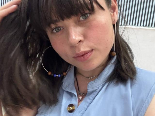 Iniridax has brown hair, blue eyes, and pink lips, wearing a blue shirt with a button, hoop earrings, and a necklace. She's touching her hair with a slight smile.