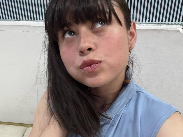 Iniridax with pouty lips, brown eyes looking up, and dark bangs, wearing a blue shirt, showing freckles and slight blush on her face, with long brown hair.
