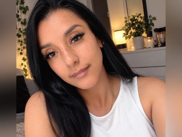 Lincysexx con capelli neri, occhi marroni, piercing al naso, lentiggini e canotta bianca, sorride alla fotocamera, ha lunghe ciglia, un sedere piccolo, luce soffusa, stanza accogliente, piante, candele sullo sfondo..