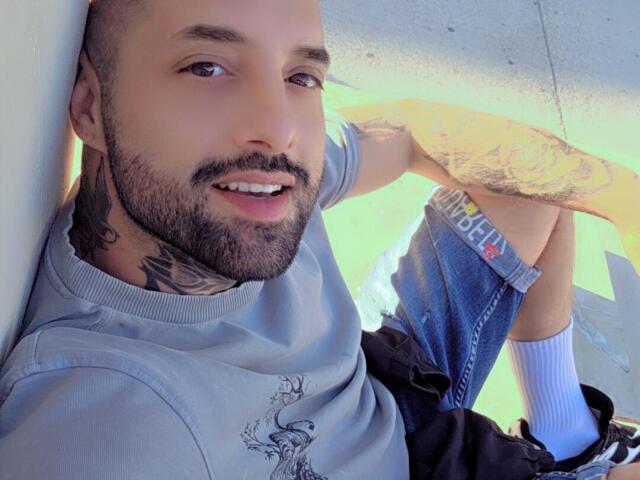 TommyDum, con la cabeza afeitada, barba, tatuajes en el cuello y el brazo, vistiendo una camisa gris, jeans azules, calcetines blancos, sonriendo, apoyado contra una pared, mostrando una actitud relajada..