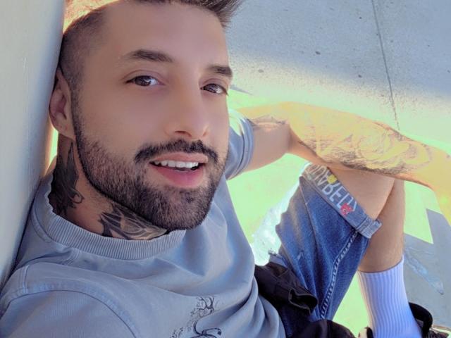 TommyDum, con barba recortada, tatuajes en el cuello y el brazo, cabello castaño corto, vistiendo una camisa gris y pantalones vaqueros azules, sonriendo, apoyado contra una pared, con las piernas cruzadas..