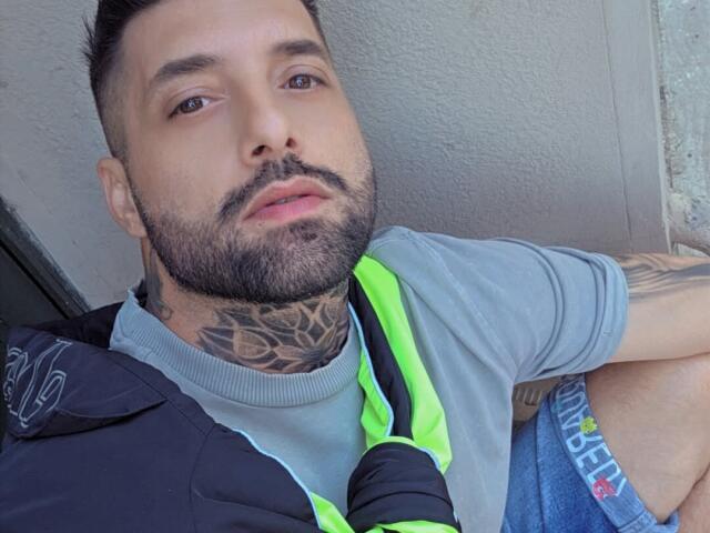 TommyDum a une barbe, des tatouages sur le cou, et porte un gilet vert néon par-dessus un t-shirt gris, avec un short en jean. Ses fesses sont partiellement visibles..