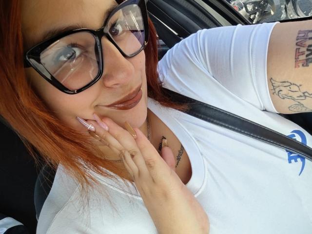 Xyrazty. Speels, met rood haar, zwarte bril en een wit shirt, die haar lip aanraakt met lange, roze nagels, waarbij tatoeages op haar arm zichtbaar zijn, zittend in een auto..