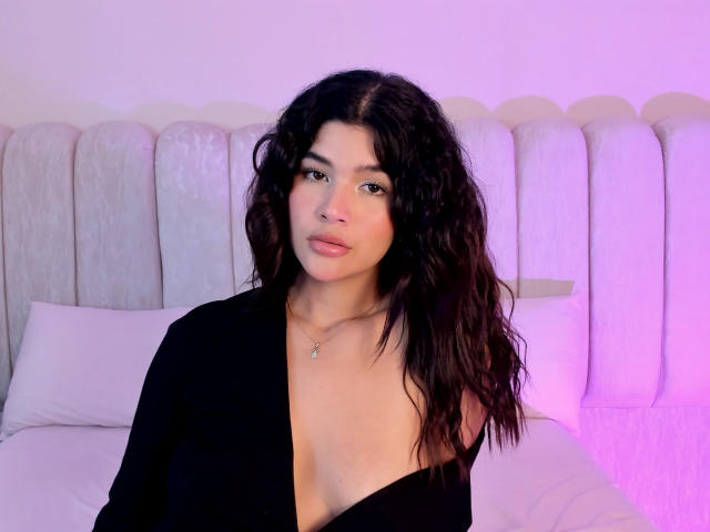 ScarlletZhen with wavy dark hair, medium tits, and a low-cut black top, wearing a necklace, against a white headboard and purple-lit background.