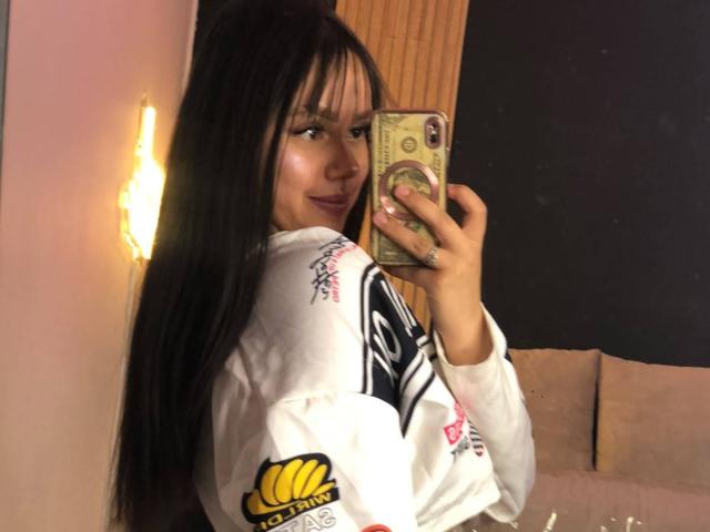MaddyHot69 with long black hair, wearing a white hoodie, taking a mirror selfie, holding a phone with a green case, looking back with a slight smile.