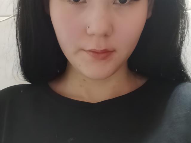 RisaDunken hat ein Gesicht mit einem kleinen Nasenring, schwarzen Haaren, heller Haut, einem leichten Rouge und einer kleinen Pickel auf dem Kinn. Sie trägt ein schwarzes Oberteil und hat ein kaum merkliches Lächeln..