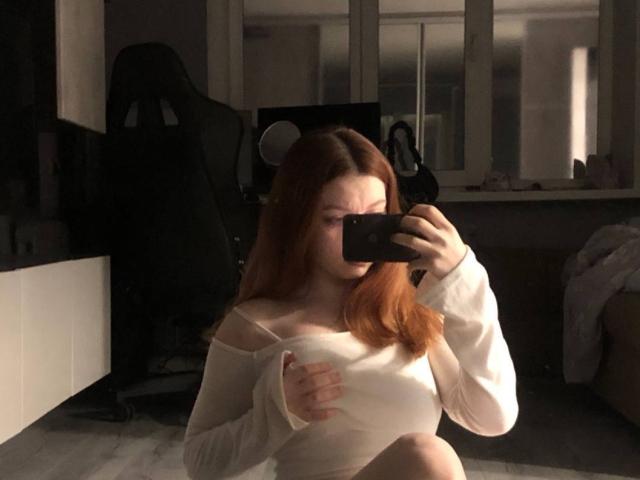 LilliamChevalier with red hair, white off-shoulder top, holding phone, covering small white tits, sitting, taking mirror selfie, dim room, black chair in background.
