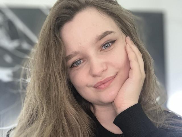 MagikPeachh, con cabello castaño largo, ojos azules y una ligera sonrisa, apoyando su mejilla en su mano, vistiendo una blusa negra, luciendo linda y relajada..