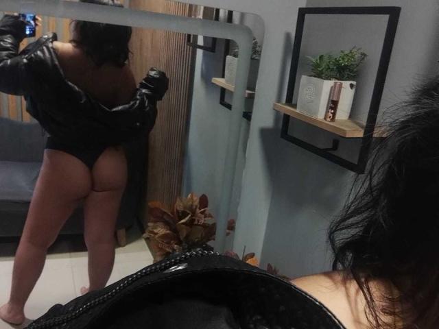 AmatistaMounX está de pie frente a un espejo, con la chaqueta de cuero negro puesta a un lado, mostrando su trasero desnudo y una tanga negra, cabello oscuro, de espaldas a la cámara, en una habitación con poca luz..