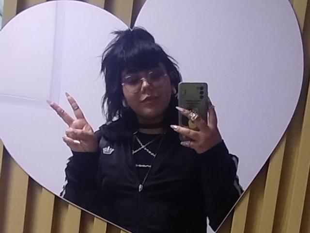 AmatistaMounX, avec ses cheveux noirs en bataille, ses lunettes et sa veste noire Adidas, fait un selfie devant un miroir en faisant un signe de la paix des deux mains, et tient un téléphone vert..