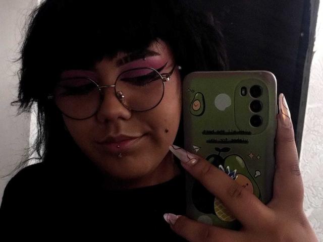 AmatistaMounX met zwart haar, roze oogschaduw, een bril, lip piercings en een zwart shirt, houdt een groene telefoon met een cartoon ontwerp vast en laat lange, verzorgde nagels zien..
