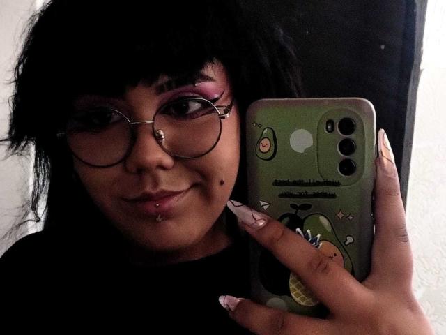 AmatistaMounX mit schwarzen Haaren, Brille, pinkem Lidschatten, Nasenring, Lippenpiercing und Septum-Piercing, hält ein grünes Telefon mit einem Cartoon-Charakter, lächelt. Dunkle, stimmungsvolle Beleuchtung..