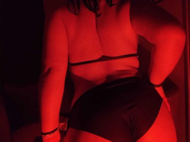 AmatistaMounX met rode verlichting, draagt een zwart, open topje en korte broek, met haar hand op haar heup, waarbij haar borsten en billen zichtbaar zijn, donker krullend haar, een armband om haar pols, en staat licht voorovergebogen..