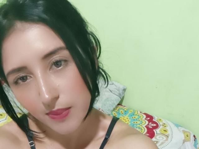 NicollJeans has short black hair, pink lips, and a slight blush on her fair skin. She's wearing a black strap, looking at the camera with a subtle smile.
