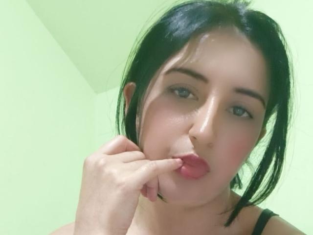 NicollJeans with black hair, pouty pink lips, and a finger in her mouth, showing her slightly flushed face and wearing a black strap. She has light skin and blue eyes.
