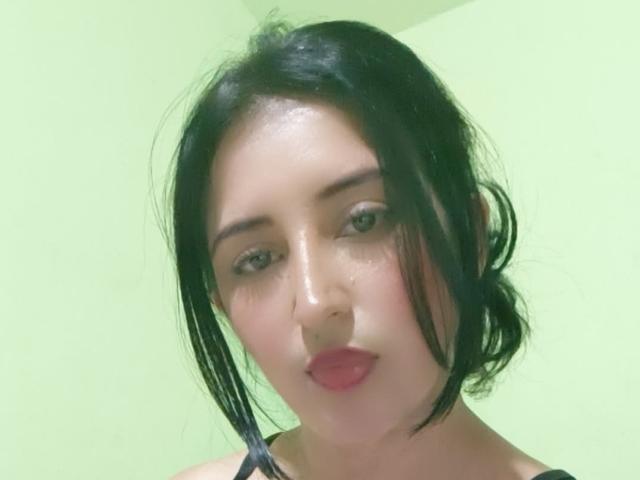 NicollJeans has black hair, light skin, and pink lipstick, with subtle makeup and a slightly sweaty face, wearing a black top, in a green-lit selfie.