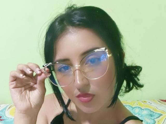 NicollJeans with black hair, wearing clear glasses, pink lipstick, black tank top, holding glasses, green wall, and colorful patterned pillow in the background.