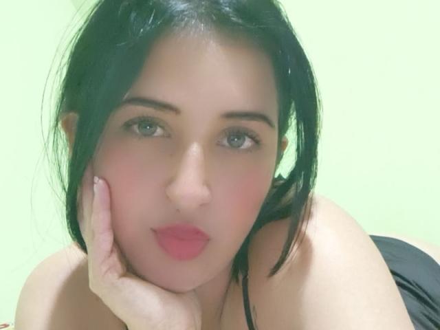NicollJeans with black hair, fair skin, and pink lips, resting her cheek on her hand, showing off her bare shoulders and a hint of black clothing.