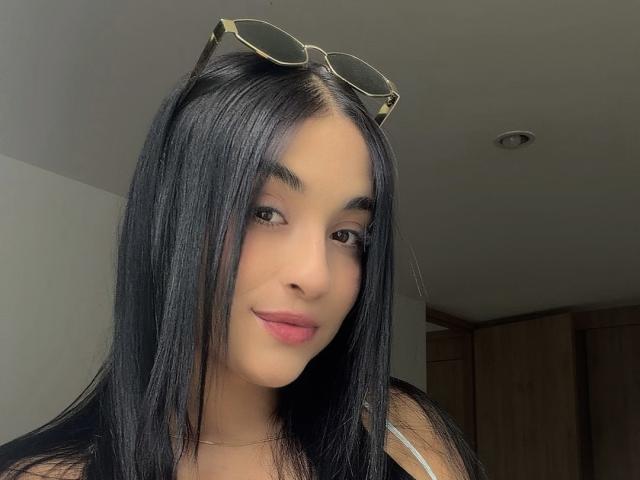 EmmaWaltter a de longs cheveux noirs, des yeux marrons et des lèvres roses. Elle porte des lunettes sur la tête et un haut noir. Ses fesses ne sont pas visibles..