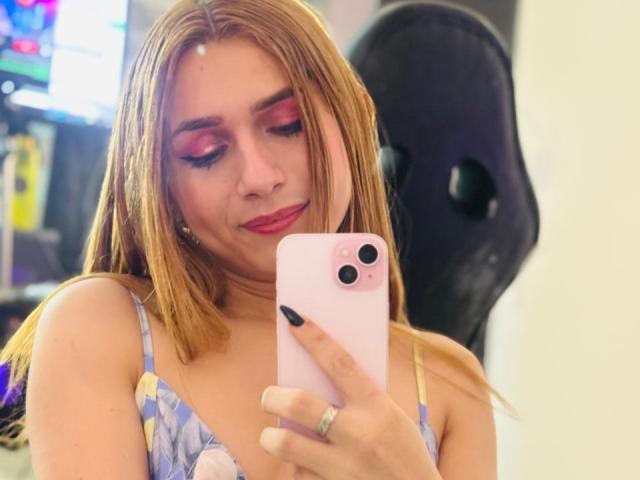 AndyDupont with blonde hair, pink eyeshadow, red lipstick, wearing a floral dress, taking a selfie with a pink iPhone, black nail polish, and a black mask in the background.