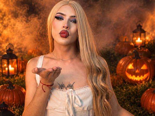 MistressNnyx, con cabello rubio largo, labios carnosos y lápiz labial rojo, vestida con un vestido blanco, mostrando sus senos con un gesto de la mano, rodeada de calabazas y faroles encendidos..