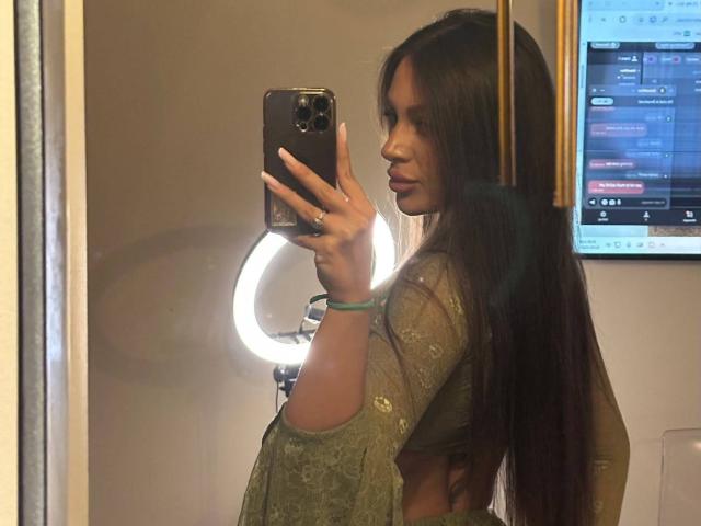 CristalSky, con lunghi capelli castani, indossa una maglietta verde, tiene il telefono per un selfie allo specchio, con una luce ad anello dietro di lei, porta un braccialetto verde, mostra una parte del seno, di fronte a uno schermo televisivo..