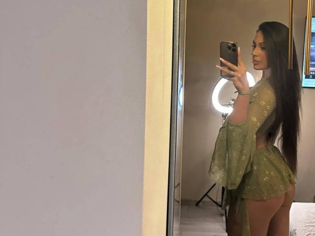 CristalSky, com cabelos castanhos longos, um vestido verde transparente e com a parte de trás do corpo à mostra, tira uma selfie em frente ao espelho, com uma luz de aro ao fundo. Ela segura um celular, olhando por cima do ombro..
