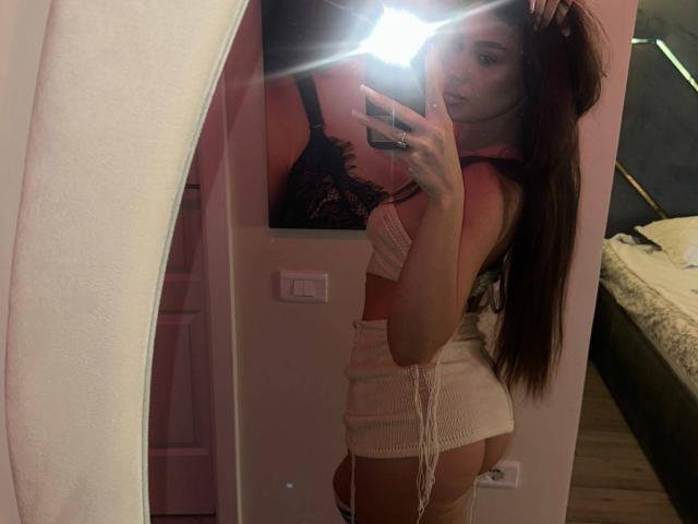 CristalSky tira uma selfie no espelho em um quarto rosa, usando um sutiã preto de renda e shorts brancos de tricô, mostrando suas nádegas e seus longos cabelos castanhos. Flash da câmera do celular..