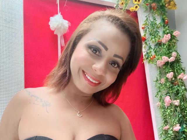 AlondraMonique with brown hair, smiling, showing tits in a black strapless top, has a tattoo on her right shoulder, wearing a necklace, and has red lipstick.