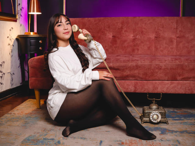 Ann Fisher, avec de longs cheveux noirs, un pull blanc, des collants noirs, tenant un téléphone vintage, assise par terre, un canapé en velours rouge derrière elle, une lampe dorée à gauche, un téléphone ancien sur le sol..