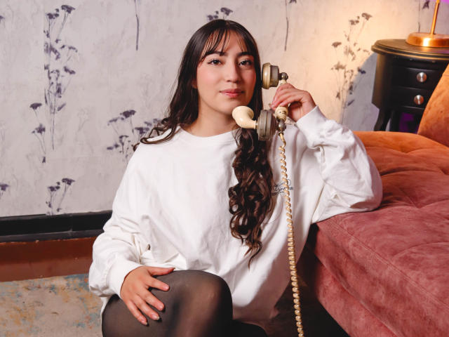 Ann Fisher, avec ses longs cheveux noirs, portant un sweat-shirt blanc et des collants noirs, assise par terre, tenant un téléphone vintage, souriant légèrement, ses fesses partiellement visibles..