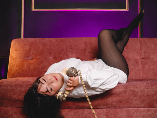 Ann Fisher, allongée sur un canapé rouge, les jambes en l'air, portant une chemise blanche et des bas noirs, tenant un téléphone d'époque, avec un éclairage violet et ses fesses bien visibles..