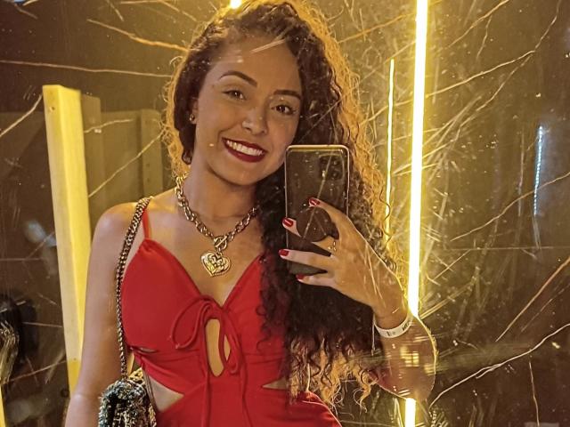 LaisMiguel with curly brown hair, red dress, heart necklace, and chain, smiling at mirror selfie, holding phone, showing red nails, and glowing yellow lights in background.